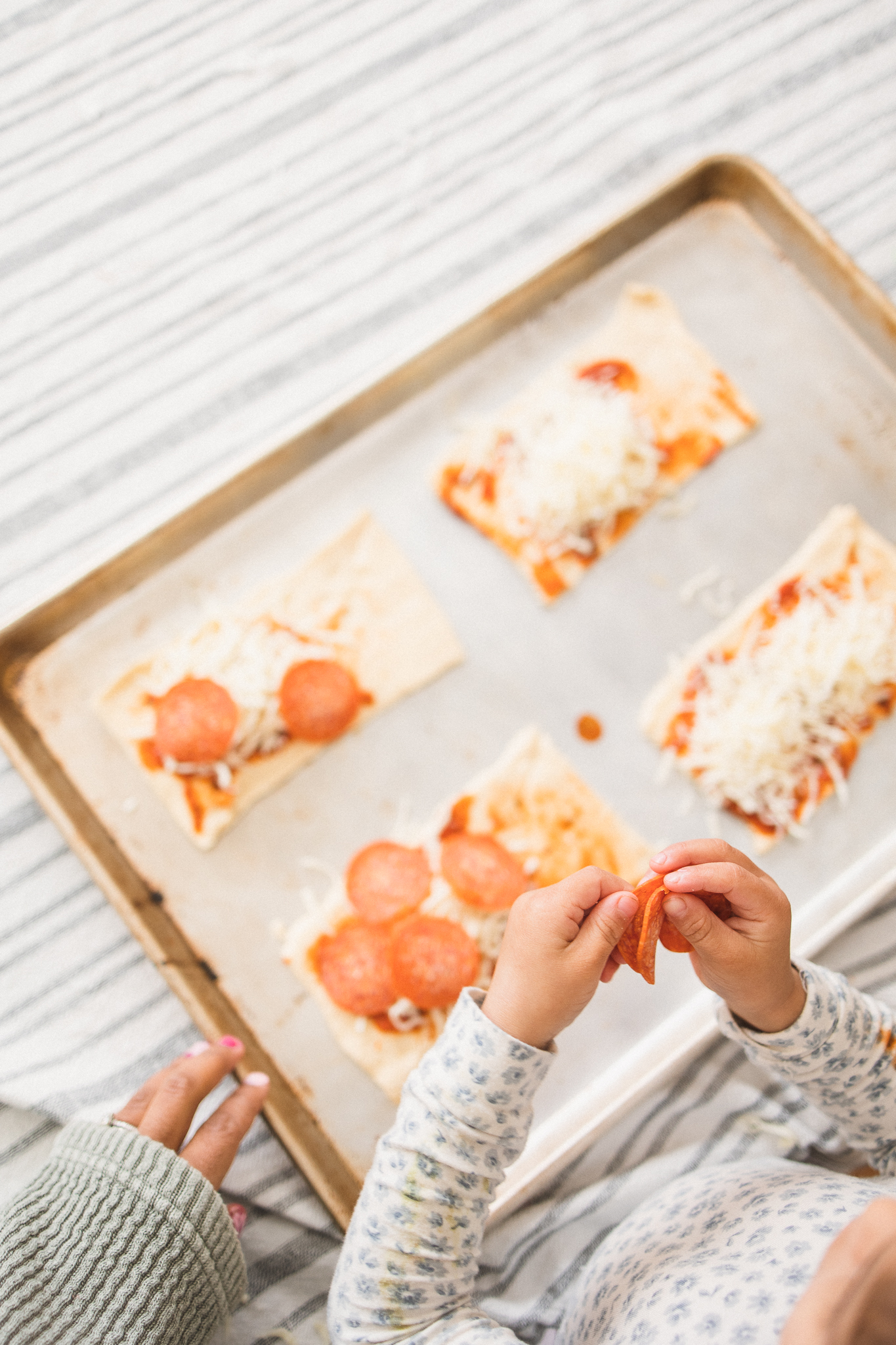 overhead shot of young child prepping pizza on a silver baking sheet with an adult hand holding the tray steady